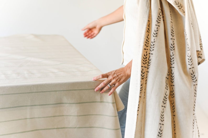 May include: A person is standing near a table with a white tablecloth with green stripes. The person is wearing a white shirt and a white shawl with a brown leaf pattern.