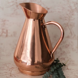 May include: A copper pitcher with a wide base and a rounded handle. The pitcher has a shiny, polished finish.