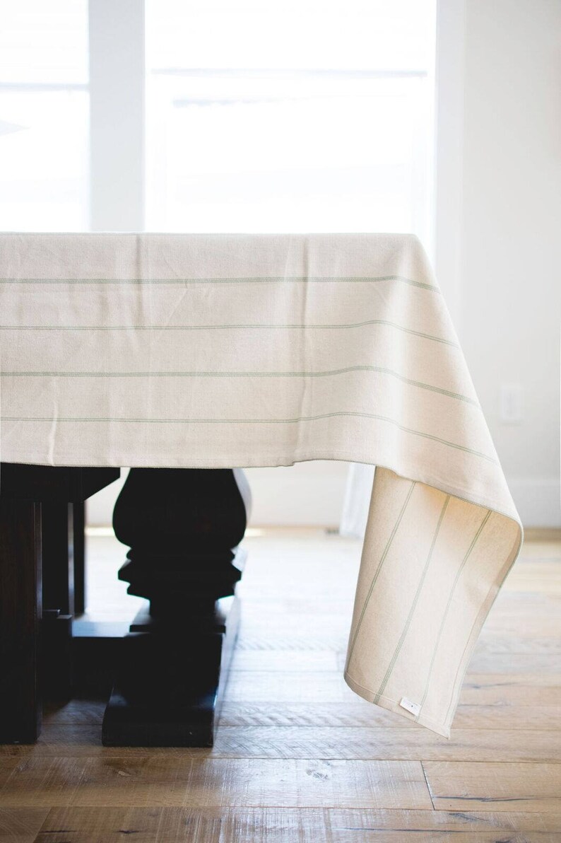 May include: A white tablecloth with thin green stripes hangs over a dark wood table. The tablecloth is draped over the table and hangs down to the floor.