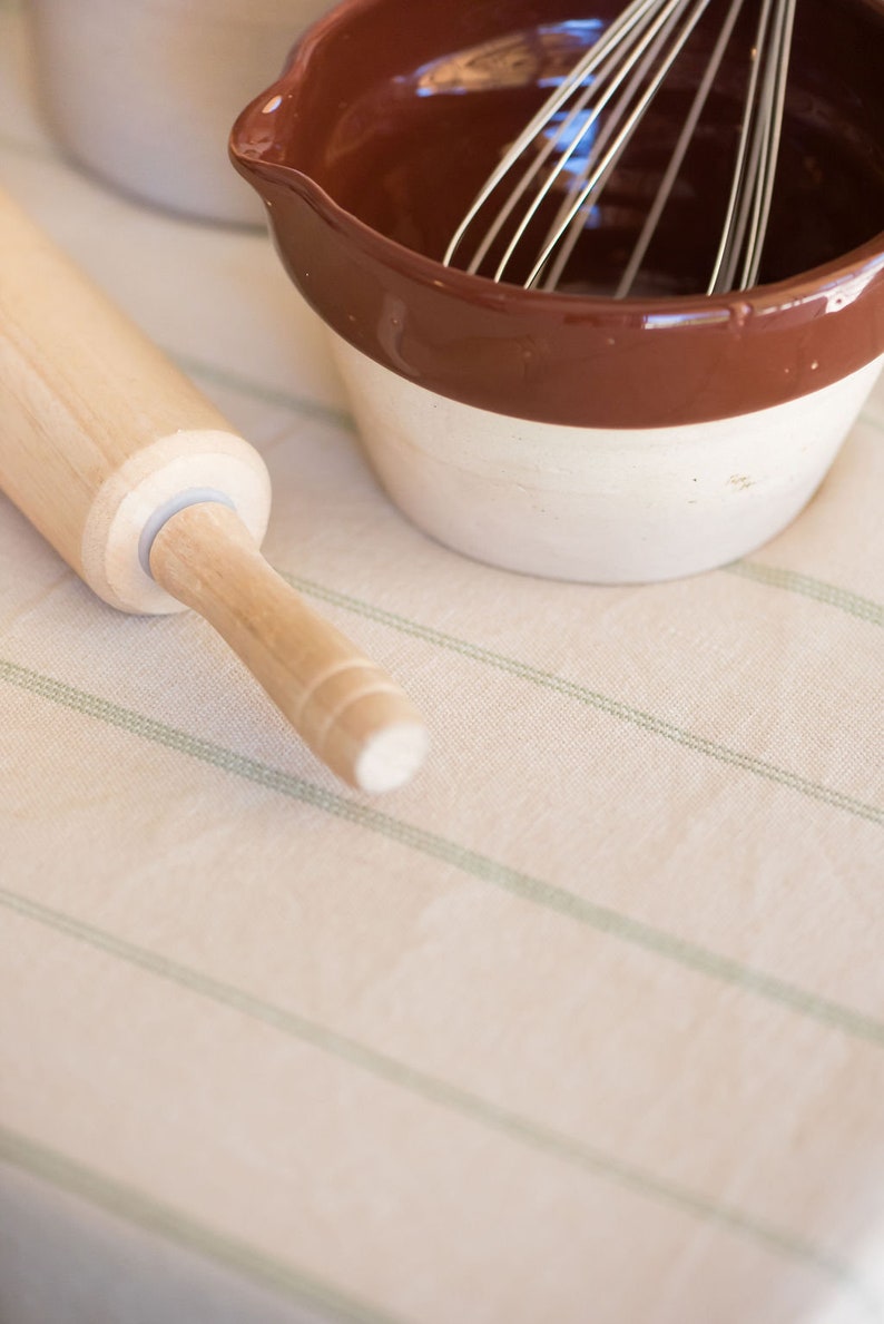 May include: A wooden rolling pin and a brown and white ceramic bowl with a wire whisk inside, on a striped tablecloth.