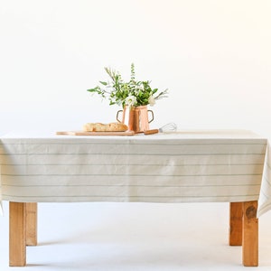 May include: A white tablecloth with thin green stripes covers a wooden table. A copper pitcher with a bouquet of white flowers and green leaves sits on the table. A wooden cutting board with a loaf of bread is also on the table.