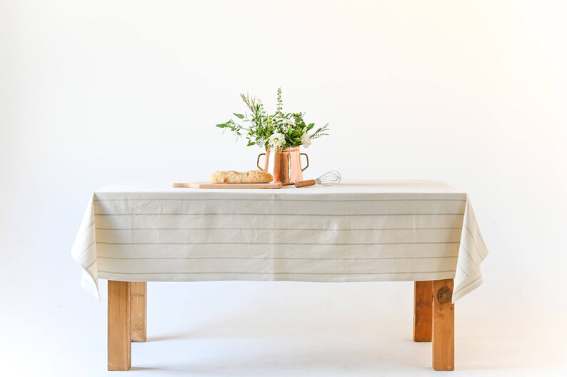 May include: A white tablecloth with thin green stripes covers a wooden table. A copper pitcher with a bouquet of white flowers and green leaves sits on the table. A wooden cutting board with a loaf of bread is also on the table.