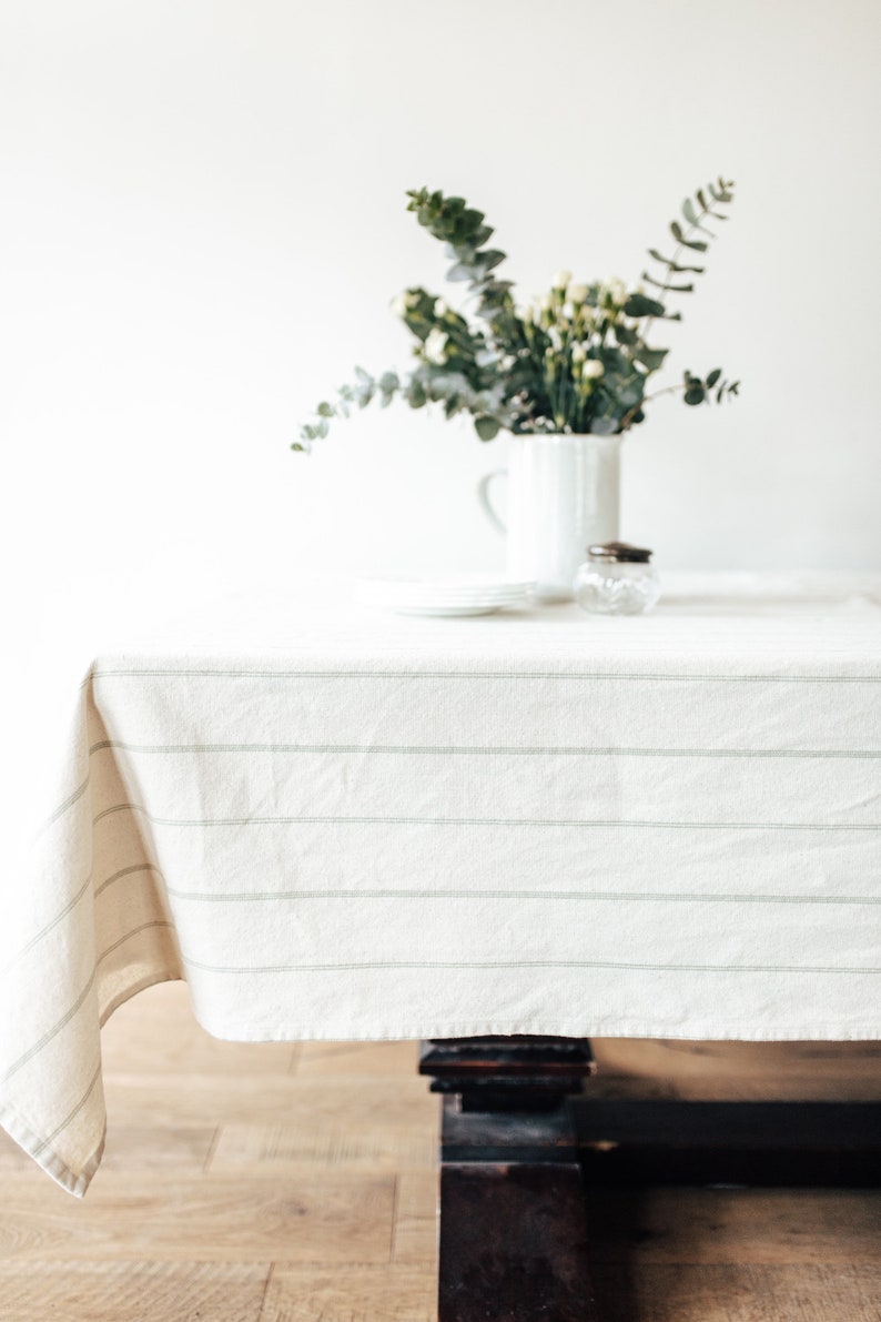May include: A white tablecloth with thin gray stripes covers a wooden table. A vase with green leaves and white flowers sits on the table.