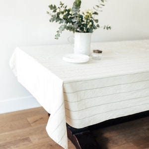 May include: A white tablecloth with thin gray stripes covers a wooden table. A white vase with a bouquet of green leaves and white flowers sits on the table.