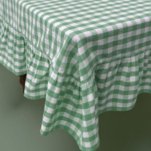 May include: A green and white checkered tablecloth with a ruffled edge. The fabric drapes over a wooden table, showcasing the classic gingham pattern. The tablecloth adds a touch of country charm to any dining setting.