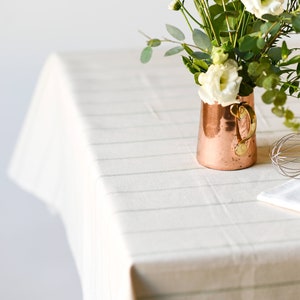 May include: A copper pitcher with a gold handle filled with white flowers and green leaves sits on a white tablecloth with a striped pattern. A silver whisk and a white kitchen towel are also on the table.