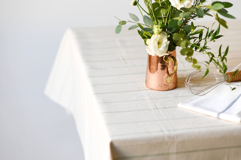 May include: A copper pitcher with a gold handle filled with white flowers and green leaves sits on a white tablecloth with a striped pattern. A silver whisk and a white kitchen towel are also on the table.