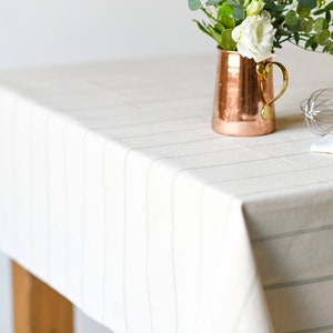 May include: A white tablecloth with thin gray stripes, a copper pitcher with white flowers, a whisk, and a white kitchen towel.