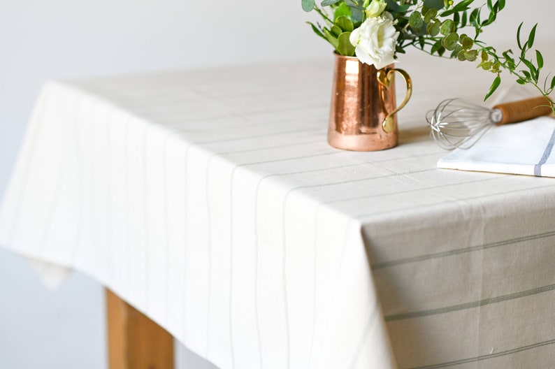 May include: A white tablecloth with thin gray stripes, a copper pitcher with white flowers, a whisk, and a white kitchen towel.