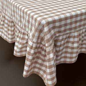 May include: A tan and white checkered tablecloth with a ruffled edge. The fabric has a classic gingham pattern, creating a rustic and inviting aesthetic. The tablecloth is draped over a wooden table.
