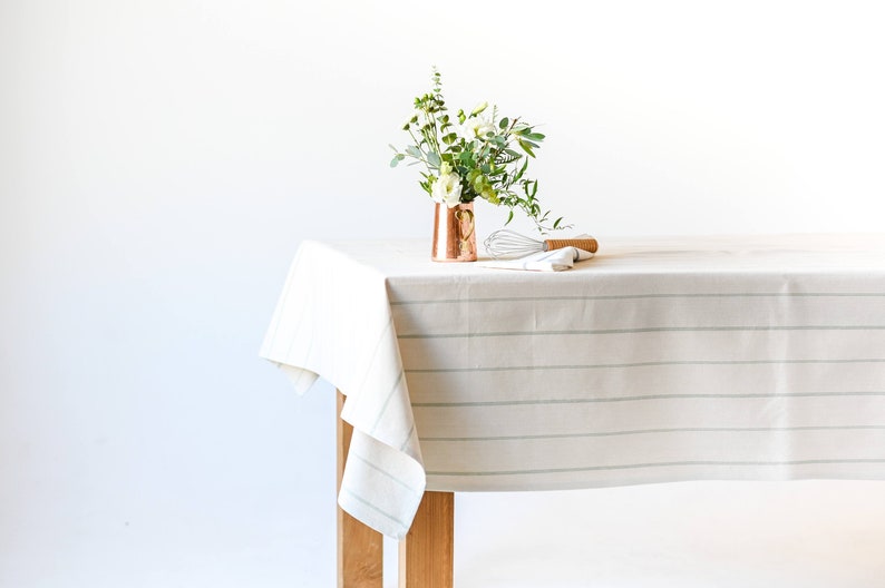 May include: A white tablecloth with thin, light green stripes, draped over a wooden table. A copper vase with a small bouquet of white flowers and greenery sits on the table, along with a whisk and a wooden utensil.