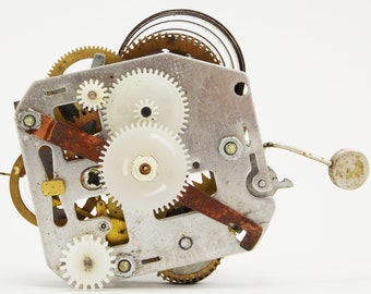 May include: Close-up of a silver and brass clockwork mechanism with multiple gears and a spring. The gears are white plastic and the spring is coiled.