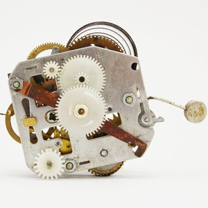 May include: Close-up of a silver and brass clockwork mechanism with multiple gears and a spring. The gears are white plastic and the spring is coiled.