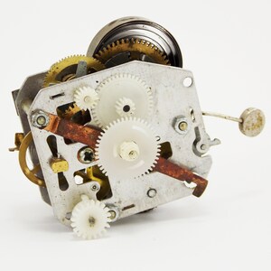May include: Close-up of a vintage clock mechanism with a variety of gears, including brass and white plastic gears. The mechanism is made of metal and has a rusty brown lever.