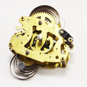 May include: Close-up of a gold-coloured clockwork mechanism with multiple gears and springs. The mechanism is disassembled and laid out on a white surface.