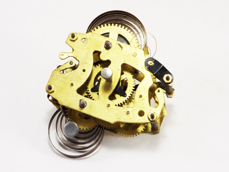 May include: Close-up of a gold-coloured clockwork mechanism with multiple gears and springs. The mechanism is disassembled and laid out on a white surface.
