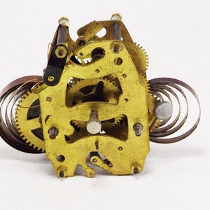 May include: Close-up of a gold-coloured clock mechanism with a coiled spring and gears. The mechanism is made of metal and has a worn, antique look.