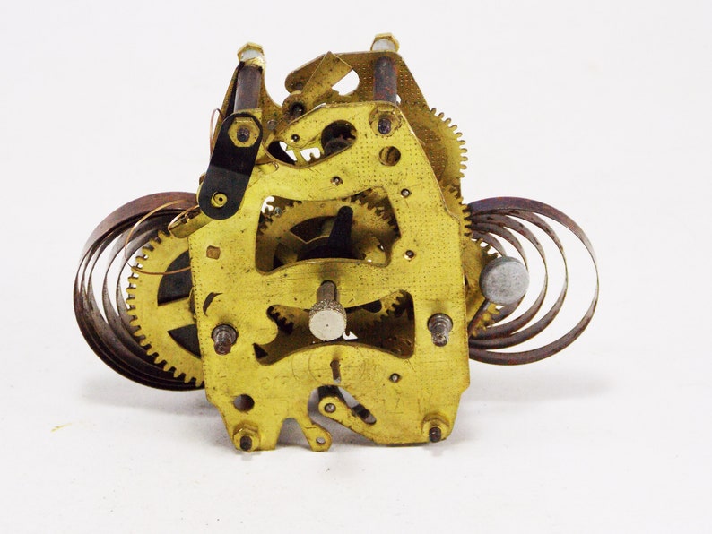 May include: Close-up of a gold-coloured clock mechanism with a coiled spring and gears. The mechanism is made of metal and has a worn, antique look.