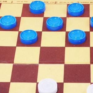 Pode incluir: Um jogo de damas em andamento com pe&ccedil;as azuis e brancas. O tabuleiro &eacute; de madeira e tem um padr&atilde;o quadriculado marrom e amarelo. As pe&ccedil;as s&atilde;o de pl&aacute;stico e t&ecirc;m uma superf&iacute;cie lisa e arredondada.