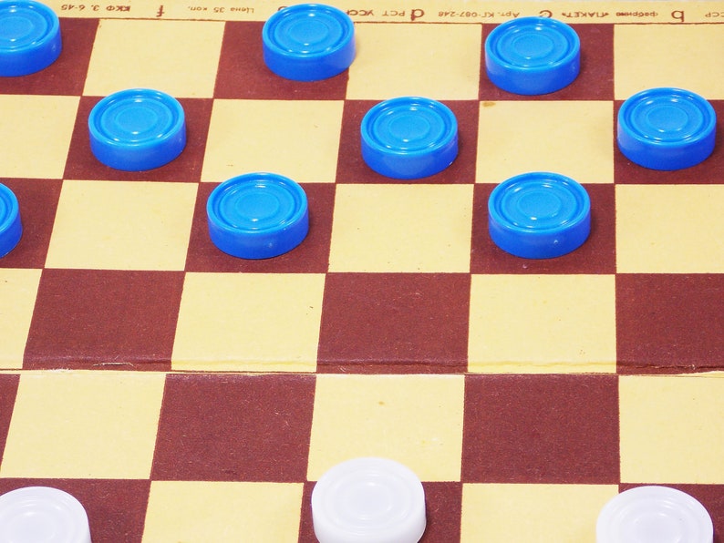 Pode incluir: Um jogo de damas em andamento com pe&ccedil;as azuis e brancas. O tabuleiro &eacute; de madeira e tem um padr&atilde;o quadriculado marrom e amarelo. As pe&ccedil;as s&atilde;o de pl&aacute;stico e t&ecirc;m uma superf&iacute;cie lisa e arredondada.