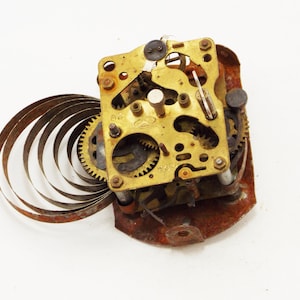 May include: Close-up of a disassembled clock mechanism. The brass-colored gears and plates are detailed, with visible screws and a rusty, reddish-brown base. A coiled metal spring extends from the mechanism, suggesting a vintage or antique item.