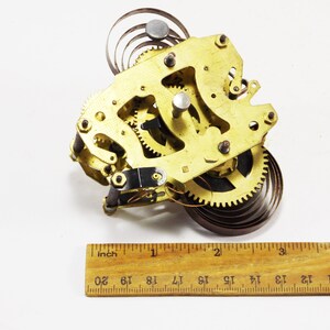 May include: A gold-coloured clock mechanism with a spring and gears. The mechanism is approximately 5 cm wide and 5 cm tall.