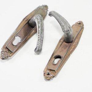 May include: Two antique door handles. The handles are silver-coloured with a detailed leaf design. The base plates are bronze-coloured with a keyhole and floral accents. The handles are likely made of metal and are for a vintage door.