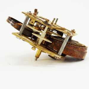May include: A rusty, antique clock mechanism with brass gears and a white plastic gear. The mechanism is missing a few parts and is in need of repair.