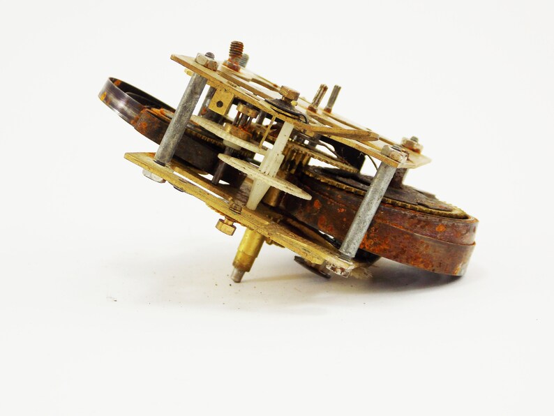 May include: A rusty, antique clock mechanism with brass gears and a white plastic gear. The mechanism is missing a few parts and is in need of repair.