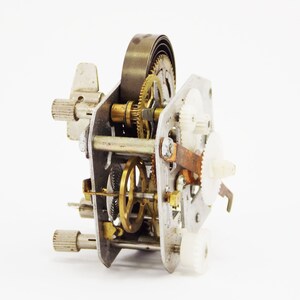 May include: Close-up of a silver and brass clockwork mechanism with gears and a spring. The mechanism is disassembled and laid out on a white surface.