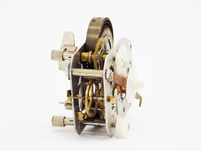 May include: Close-up of a silver and brass clockwork mechanism with gears and a spring. The mechanism is disassembled and laid out on a white surface.