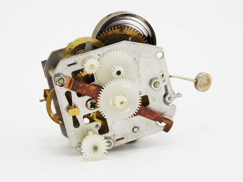 May include: Close-up of a vintage clock mechanism with a variety of gears, including brass and white plastic gears. The mechanism is made of metal and has a rusty brown lever.