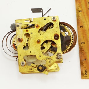 May include: Close-up of a brass clockwork mechanism with multiple gears and a spring. The mechanism is approximately 2 inches wide and 2 inches tall. The numbers "6973" and "68-1" are visible on the mechanism.
