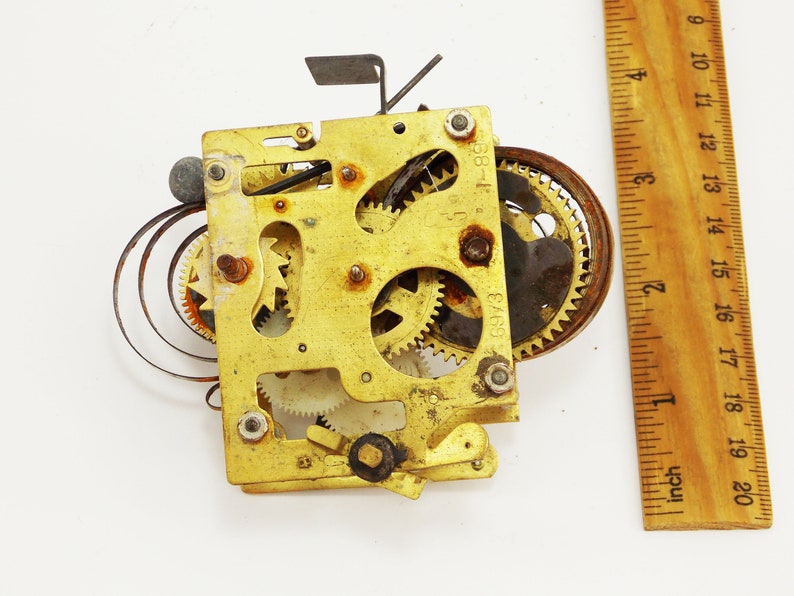 May include: Close-up of a brass clockwork mechanism with multiple gears and a spring. The mechanism is approximately 2 inches wide and 2 inches tall. The numbers "6973" and "68-1" are visible on the mechanism.