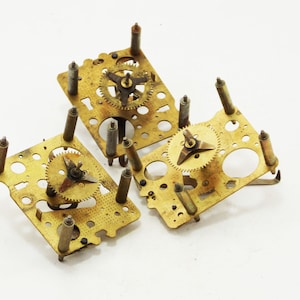 Op de afbeelding: Drie goudkleurige metalen klokveren met drie-armige armen. De veren zijn bevestigd aan metalen platen met gaten en zijn in een driehoekige vorm gerangschikt.