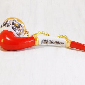 May include: A red and white ceramic pipe with a gold chain. The pipe is decorated with floral designs.
