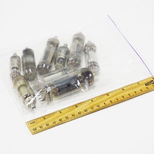 May include: A clear plastic bag containing a collection of vintage vacuum tubes. The tubes are of various sizes and shapes, with glass and metal components. A wooden ruler is placed next to the bag for scale.