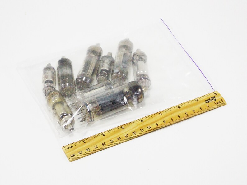 May include: A clear plastic bag containing a collection of vintage vacuum tubes. The tubes are of various sizes and shapes, with glass and metal components. A wooden ruler is placed next to the bag for scale.
