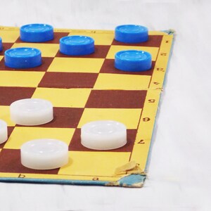 Pode incluir: Um jogo de damas com pe&ccedil;as azuis e brancas. O tabuleiro &eacute; feito de papel&atilde;o e tem um padr&atilde;o quadriculado marrom e amarelo. Os n&uacute;meros 1, 2, 6, 7, 8 e 9 s&atilde;o vis&iacute;veis na lateral do tabuleiro.