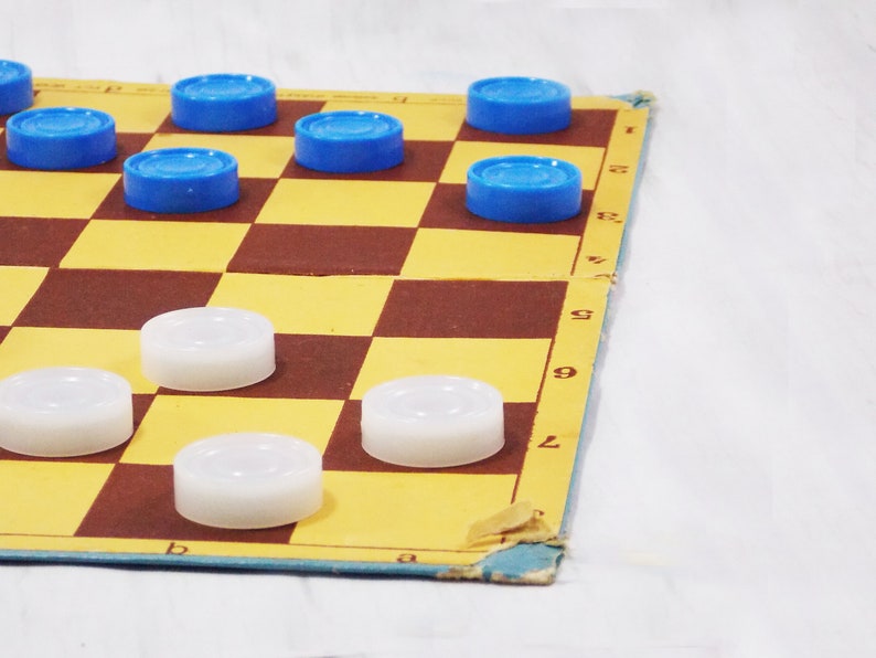 Pode incluir: Um jogo de damas com pe&ccedil;as azuis e brancas. O tabuleiro &eacute; feito de papel&atilde;o e tem um padr&atilde;o quadriculado marrom e amarelo. Os n&uacute;meros 1, 2, 6, 7, 8 e 9 s&atilde;o vis&iacute;veis na lateral do tabuleiro.