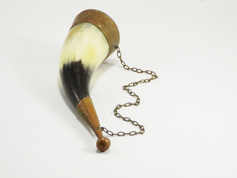May include: A brown and white cow horn drinking vessel with a brass rim and a brass chain attached to a brass ball at the end.