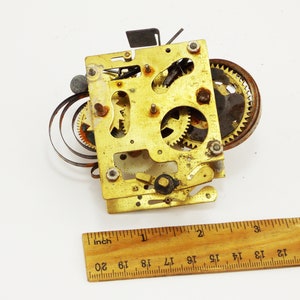 May include: Close-up of a gold-colored clock mechanism with a spring and gears. The mechanism is about 2 inches wide.