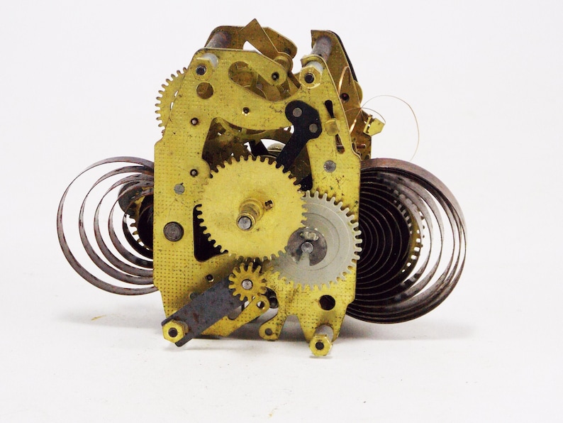 May include: Close-up of a gold-toned clock mechanism with a spring and gears. The mechanism is disassembled and laid out on a white surface.