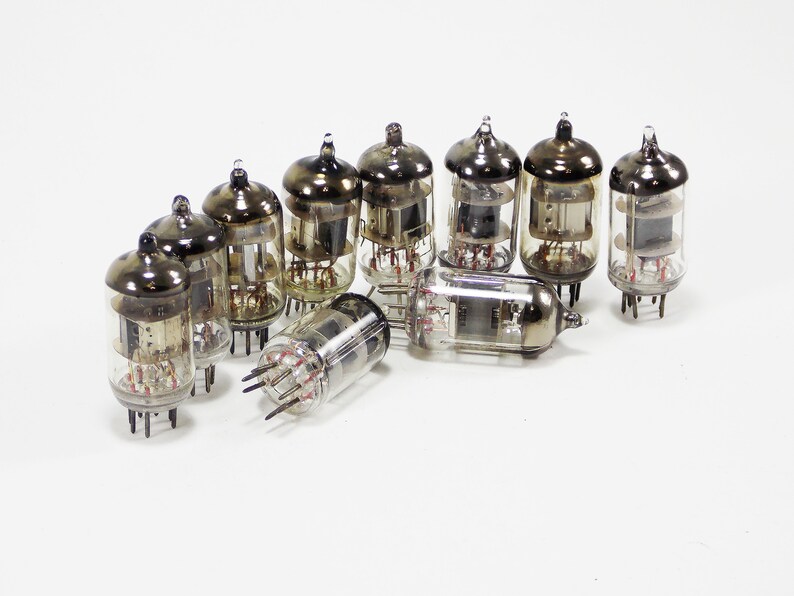 May include: A group of ten vintage vacuum tubes with glass enclosures and metal bases. The tubes are arranged in a semi-circle on a white surface.