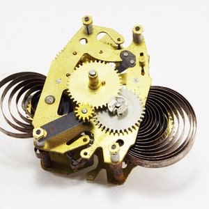 May include: Close-up of a gold-toned clockwork mechanism with multiple gears and a coiled spring. The mechanism is disassembled and laid out on a white surface.