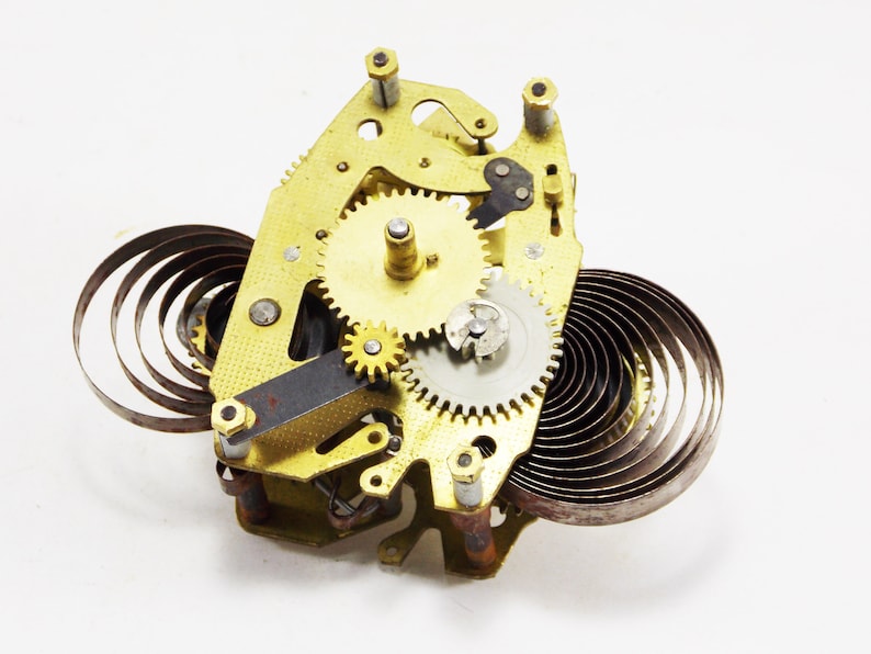 May include: Close-up of a gold-toned clockwork mechanism with multiple gears and a coiled spring. The mechanism is disassembled and laid out on a white surface.