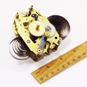 May include: Close-up of a gold-coloured clockwork mechanism with a coiled spring. The mechanism is about 5 cm wide and 5 cm tall.