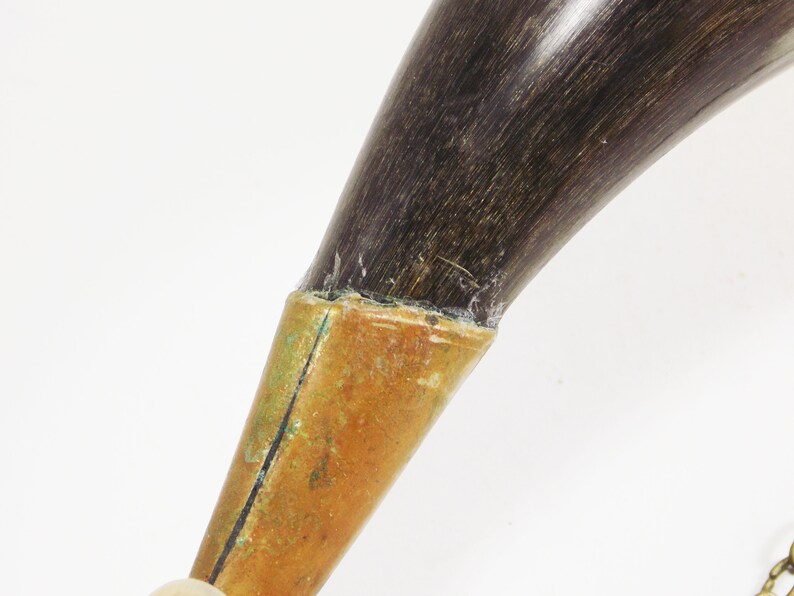 May include: A dark brown powder horn with a brass tip and a chain attached. The horn is made of natural material and has a rustic look.