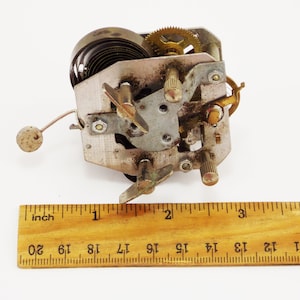 May include: Close-up of a silver and brass clockwork mechanism with a spring, gears, and a lever. The mechanism is about 2.5 cm wide and is sitting on a white surface next to a wooden ruler.