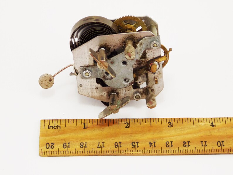May include: Close-up of a silver and brass clockwork mechanism with a spring, gears, and a lever. The mechanism is about 2.5 cm wide and is sitting on a white surface next to a wooden ruler.